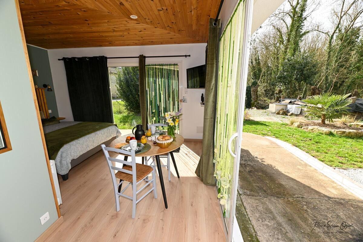 A room with a bed, a dining table with chairs, a small kitchen area, and a view of the garden.