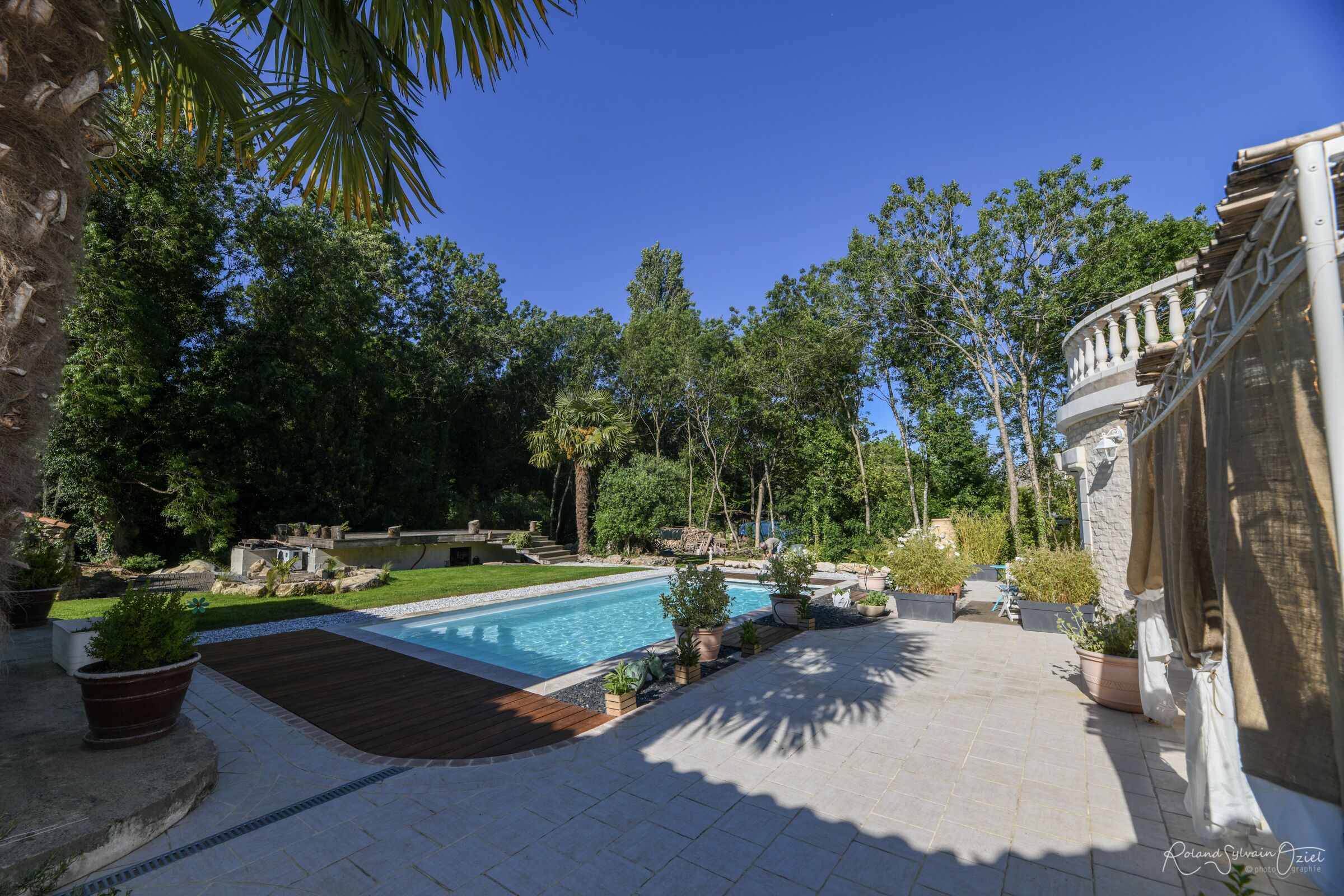 Une piscine entourée d’un jardin aménagé, avec un mur en pierre et une terrasse en bois.