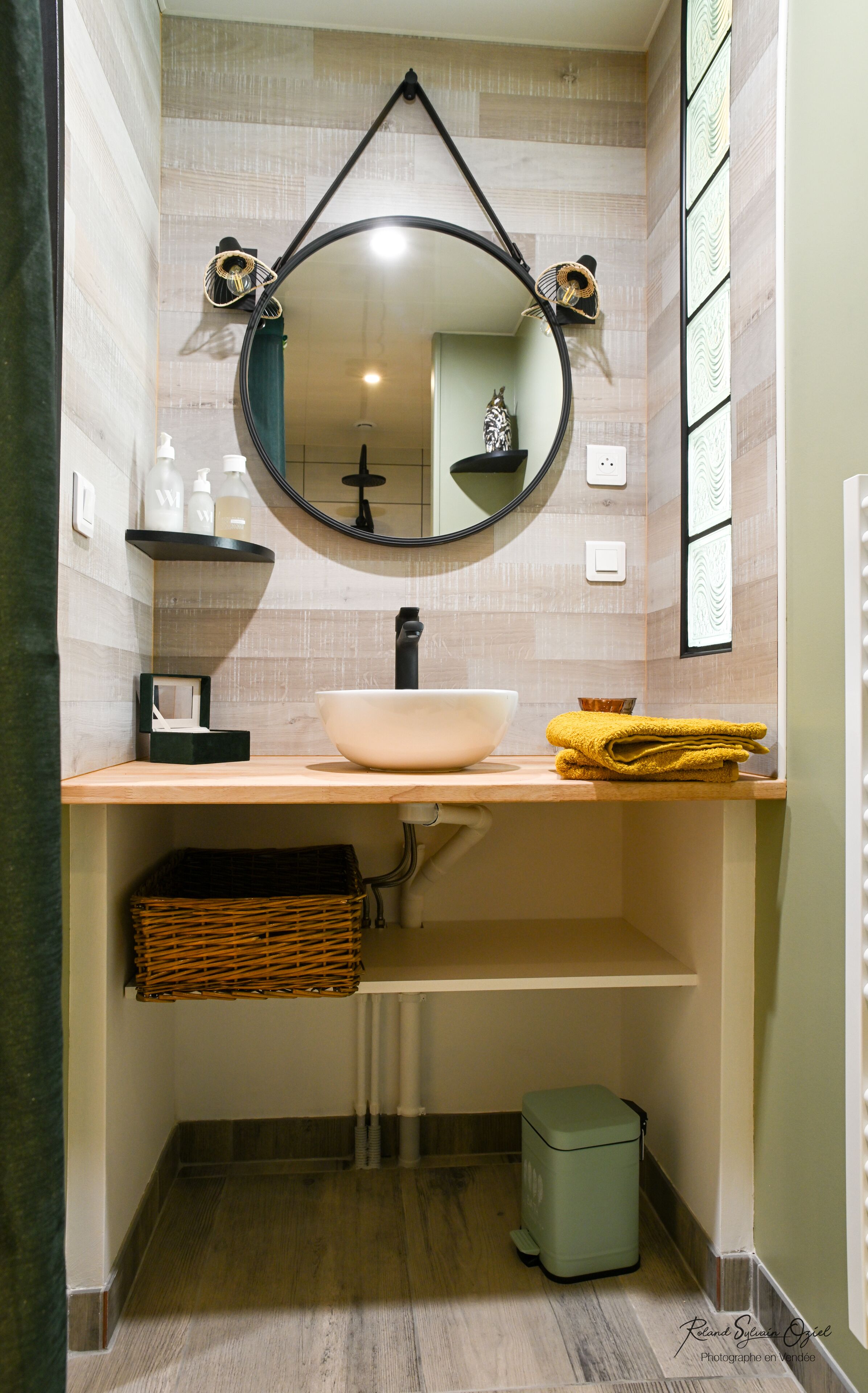 Een badkamer met een ronde spiegel, een witte wastafel, een handdoekhouder en een kleine prullenbak.