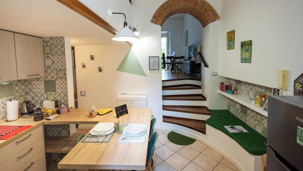 Cocina compacta con una mesa de madera, sillas y un refrigerador. Una escalera con un arco de ladrillo conduce a otra habitación.