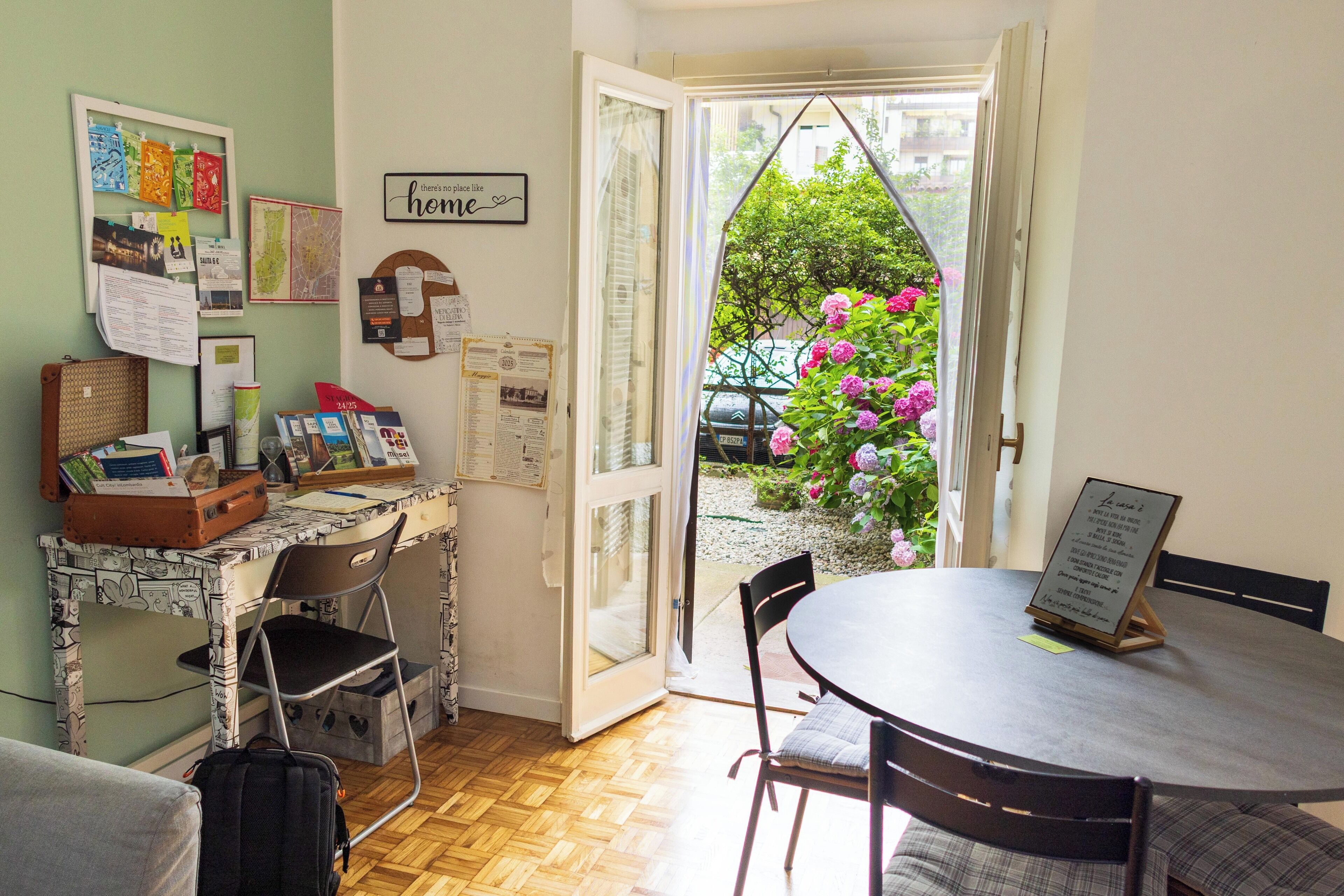 Een kamer met een bureau, stoel en tafel, een koffer, en uitzicht op bloemen door een open deur.
