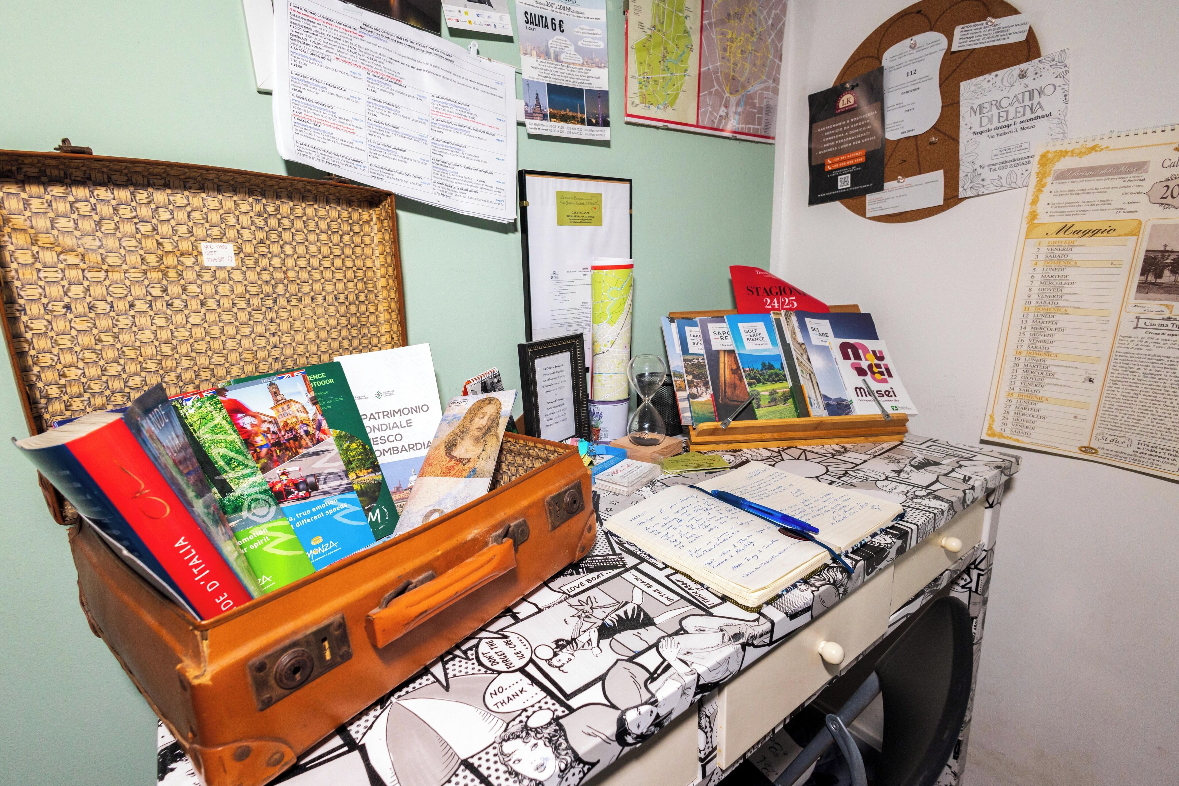 A desk with a suitcase containing books, a notepad, and pens. There are papers and a corkboard on the wall behind.
