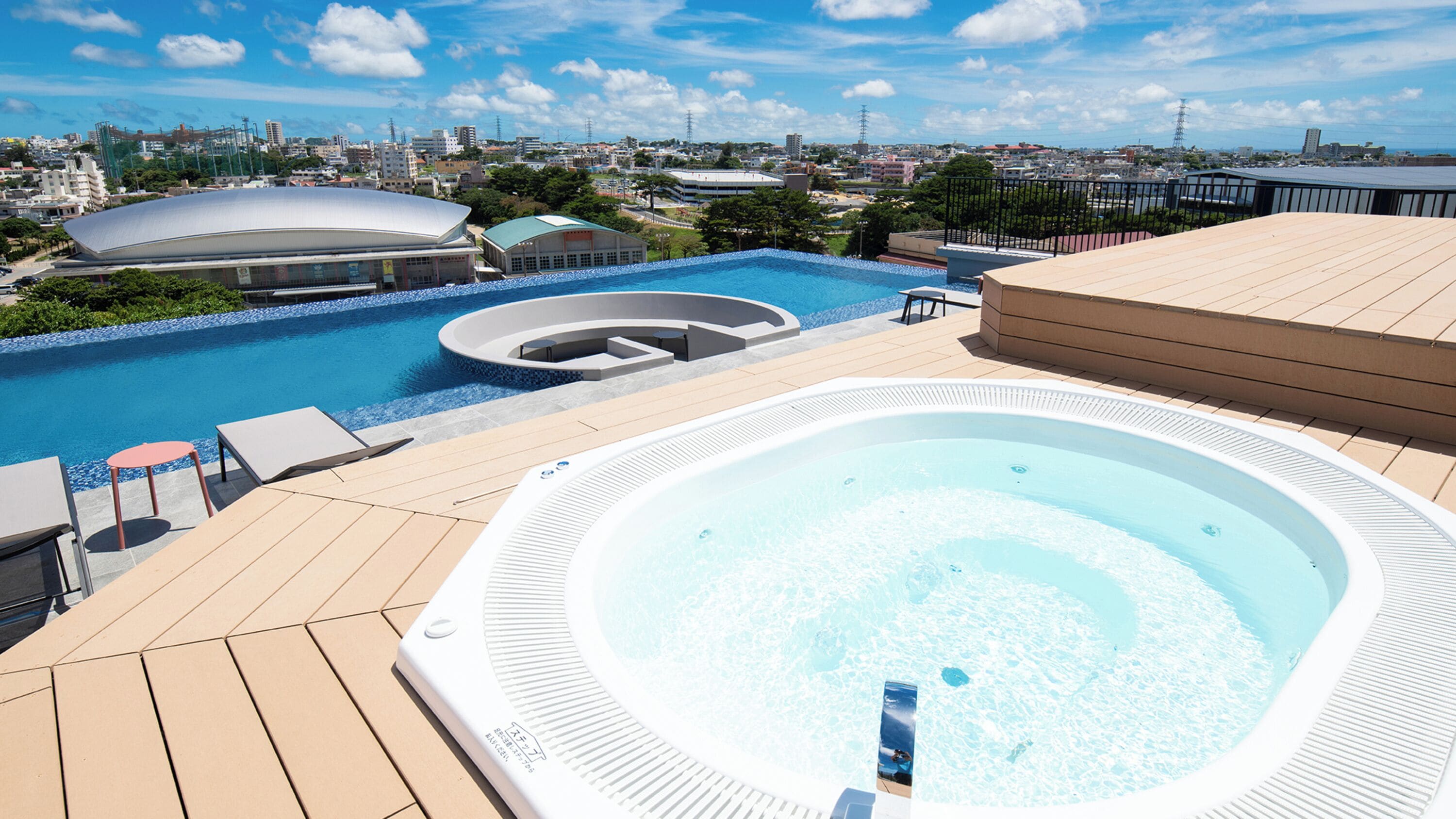 A rooftop pool area with a hot tub, lounge chairs, and a view of the cityscape.