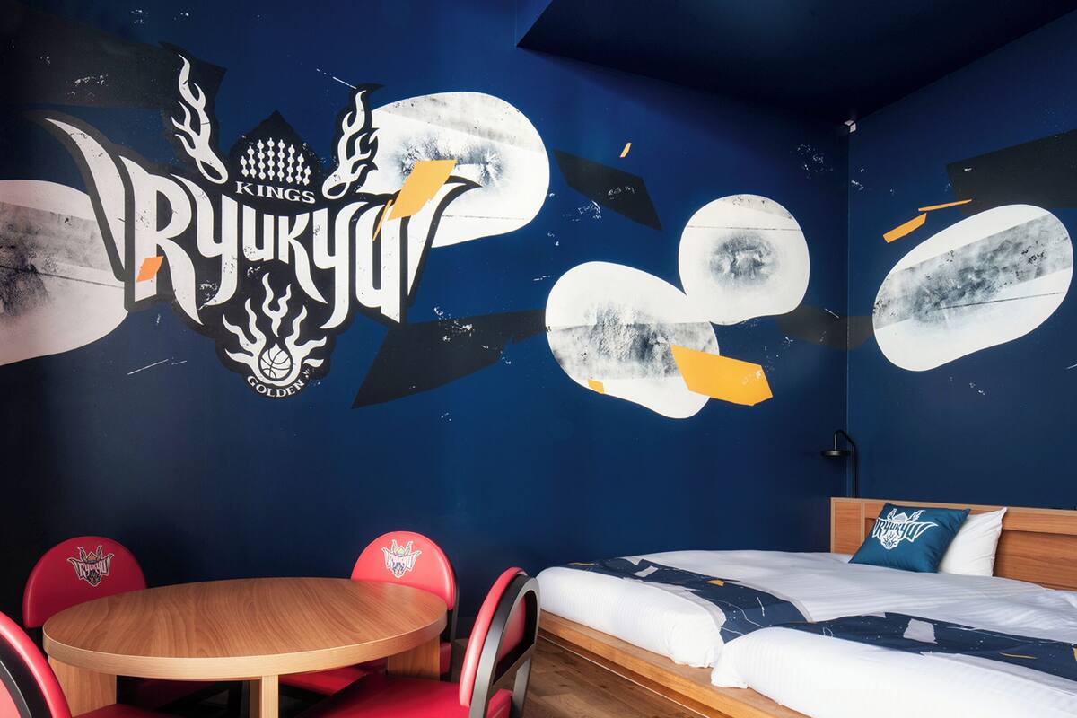 A room with a bed, a round wooden table, and red chairs. The wall features a large mural with the text "RYUKYU" and a basketball.