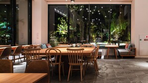 A modern restaurant with wooden tables and chairs, large windows with a view of greenery, and a seating area with cushions. - REF Okinawa Arena by VESSEL HOTELS (Okinawa)