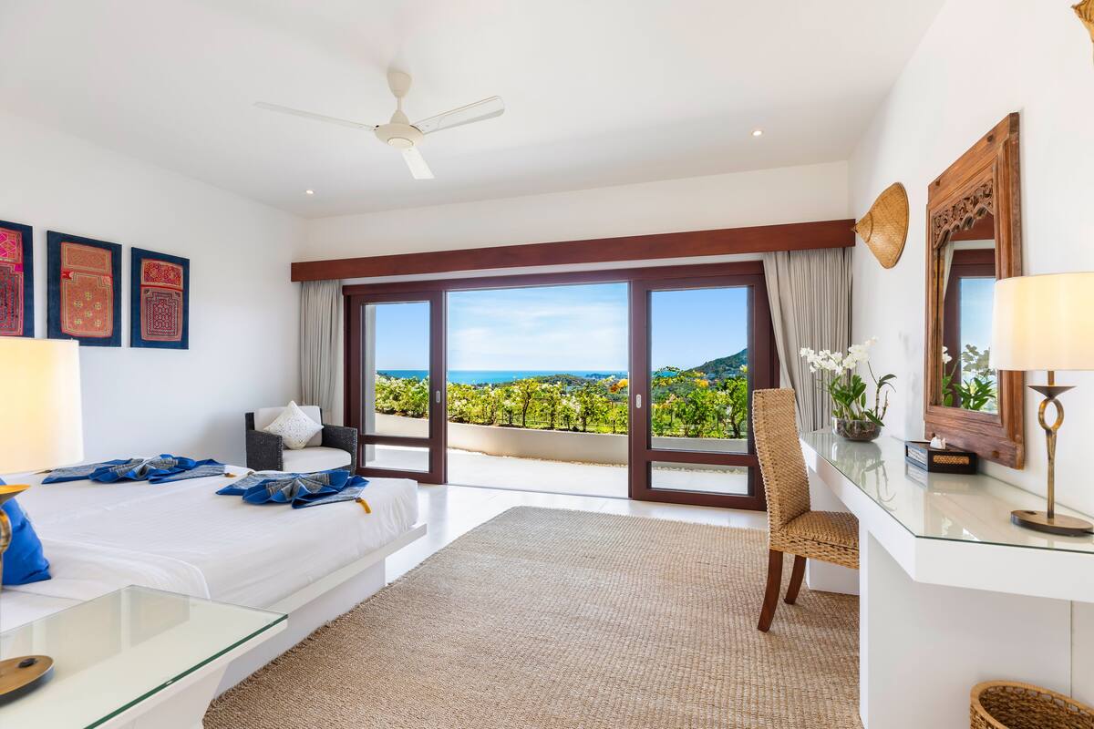 A hotel room with a large bed, a glass desk, a chair, a ceiling fan, and a view of a vineyard through large windows.