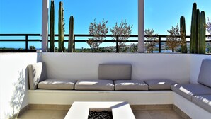 Un espace extérieur aménagé avec des sièges blancs, comprenant un banc rembourré et une petite table, le tout entouré de grands cactus et surplombé par un ciel bleu clair.