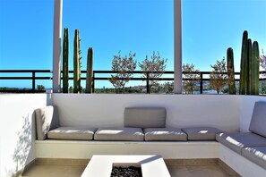 A white outdoor seating area with a cushioned bench and a small table, surrounded by tall cacti and a clear blue sky.
