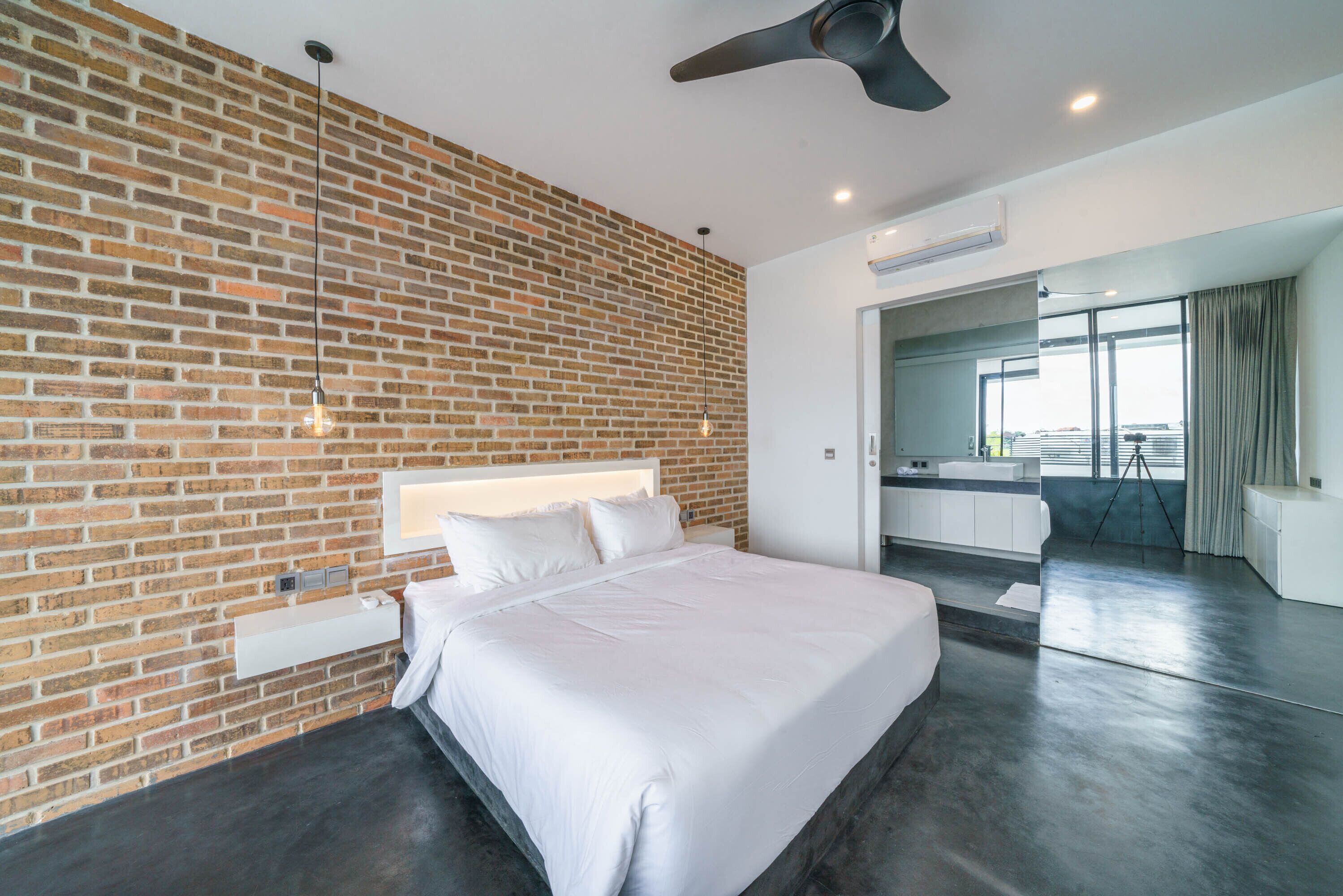 A modern hotel room with a brick wall, a large bed, a ceiling fan, and a bathroom visible through an open door.