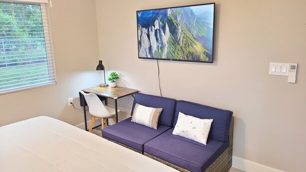 A room with a bed, a purple sofa, a desk with a lamp, a potted plant, and a wall-mounted TV displaying a mountainous landscape. - Air Conditioned 2 Bedrooms 1 Bath Apartment, fine decorated, Free Laundry in HPP (Keaau)