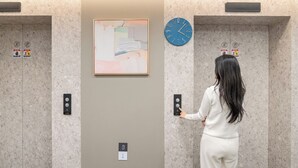 Una mujer con un conjunto blanco usando un panel de control de ascensor, con un reloj y un cuadro abstracto en la pared.