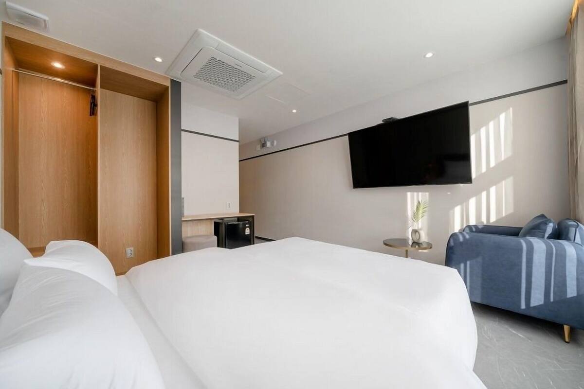 Una habitación de hotel moderna con una cama grande, un televisor en la pared, una zona de estar con sofá y un armario de madera.