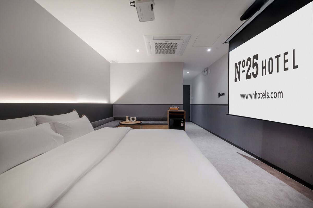 Una habitación de hotel moderna con una cama grande, una mesita de noche pequeña, una pantalla de proyector montada en la pared que muestra 'N°25 HOTEL', y una dirección web visible.