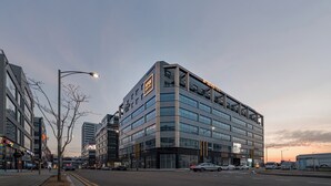 Un hotel de varios pisos con un diseño moderno, que cuenta con un letrero llamativo, ventanas grandes y una ubicación en esquina en una intersección urbana.