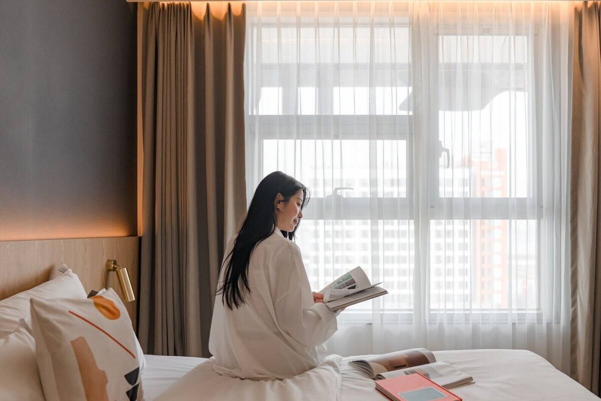 Una persona leyendo un libro en una cama en una habitación con un gran ventanal y cortinas.