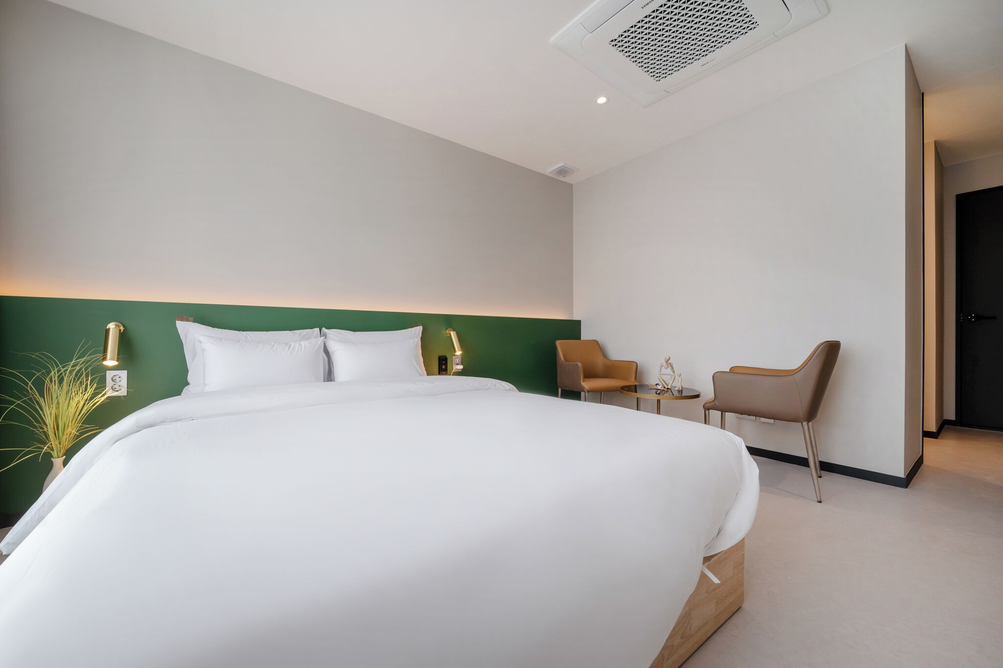 A modern hotel room with a large bed, two chairs, a small table, and a green accent wall.