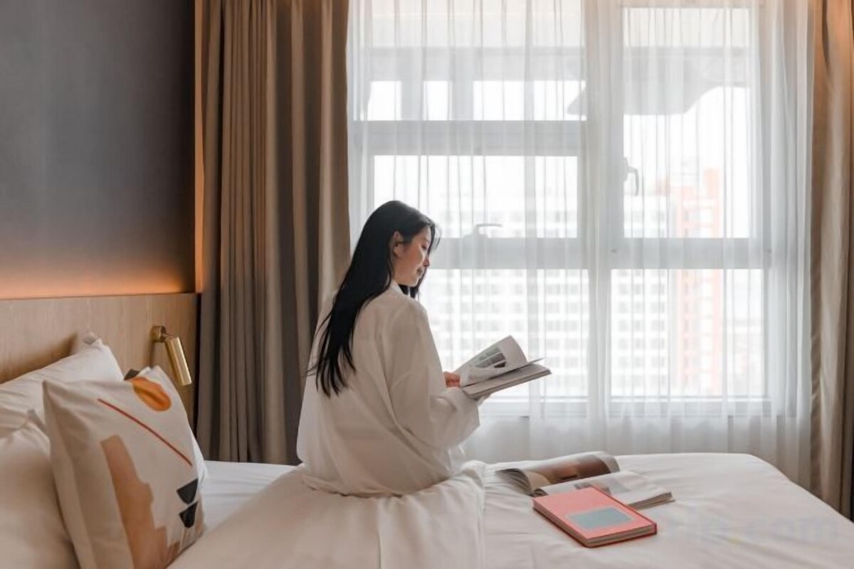 Una persona sentada en una cama en una habitación con un ventanal grande, leyendo un libro.