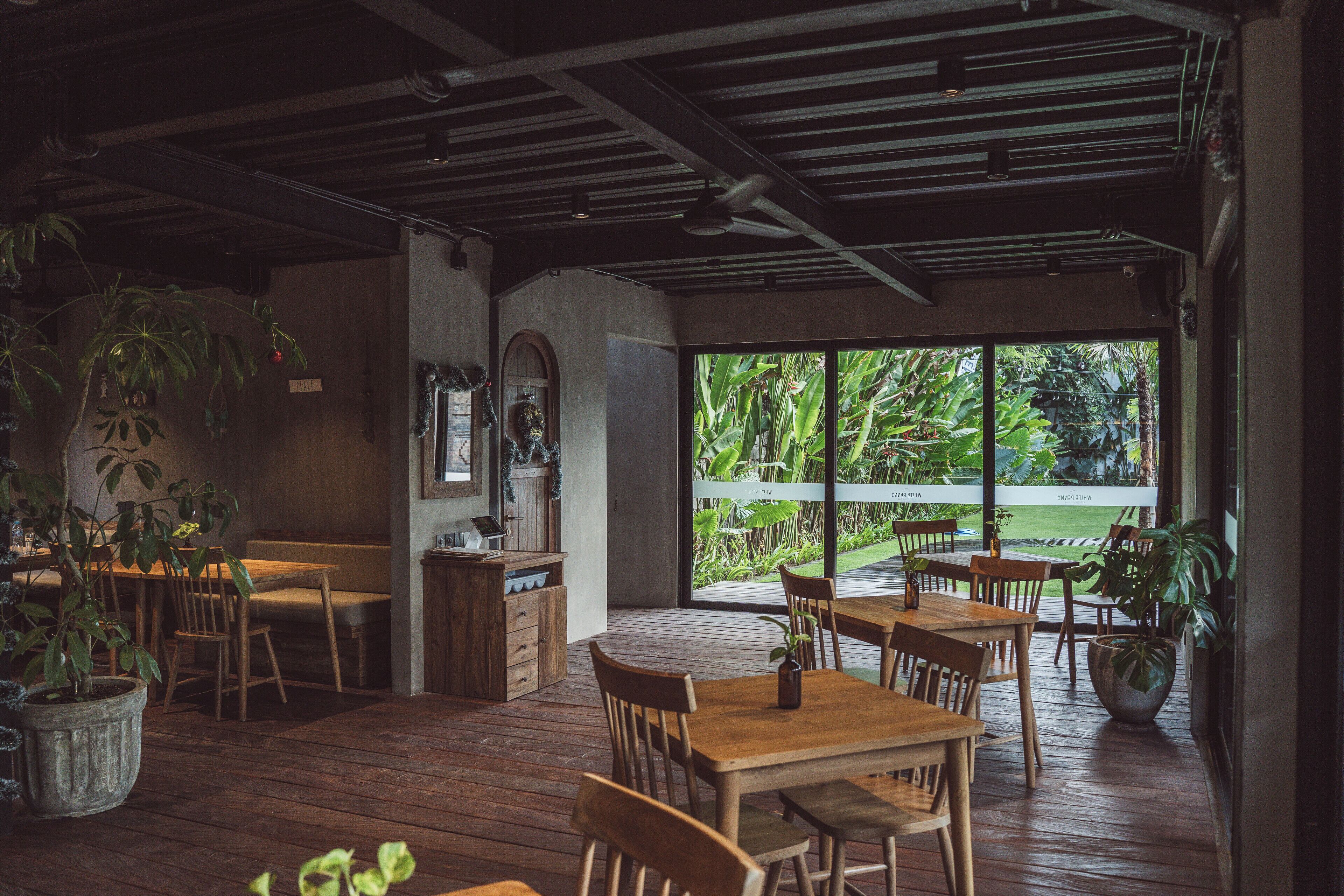 Un restaurante con mobiliario de madera, ventanas amplias y plantas en macetas.