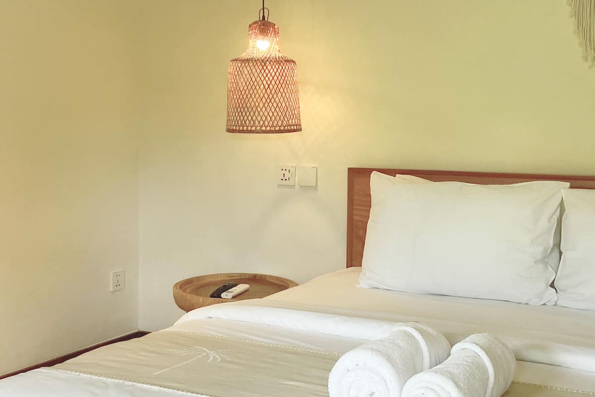 A neatly made bed with white linens and two rolled towels, a wooden bedside table with a remote control, and a hanging light fixture.