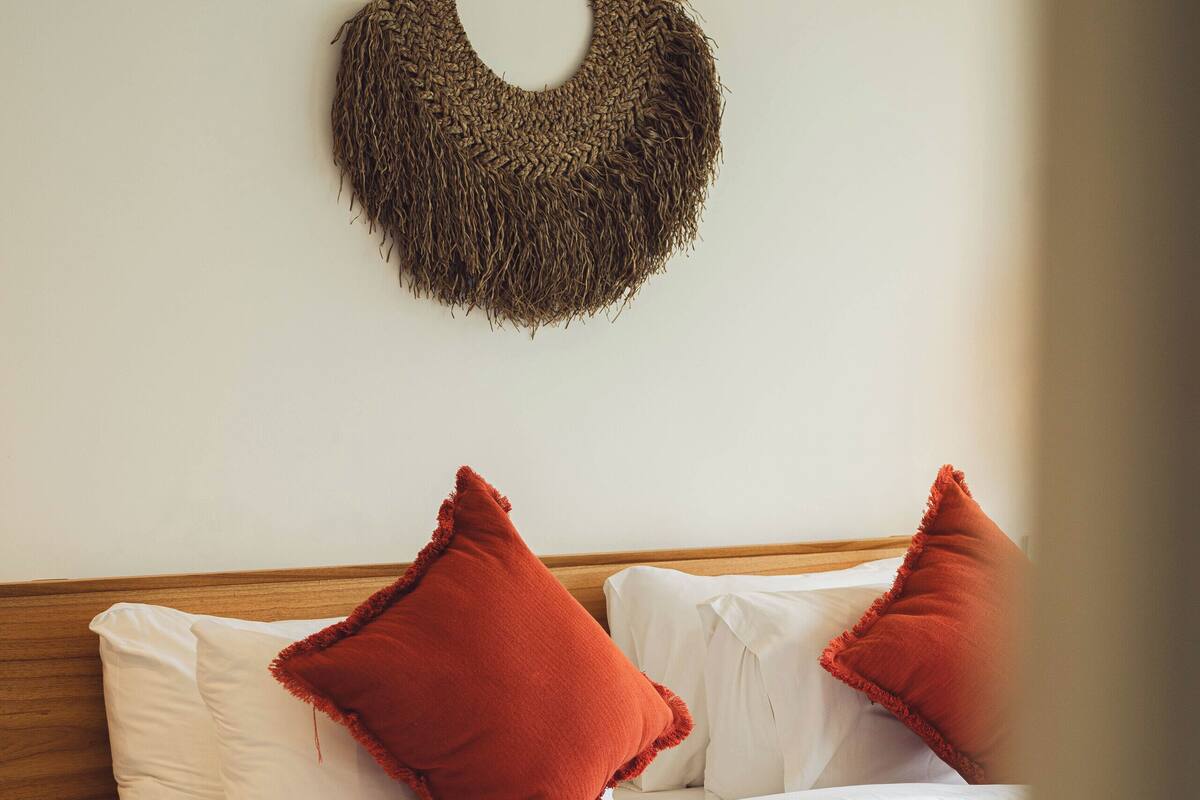 A bed with white bedding, two red pillows, a green throw blanket, and a woven wall hanging.