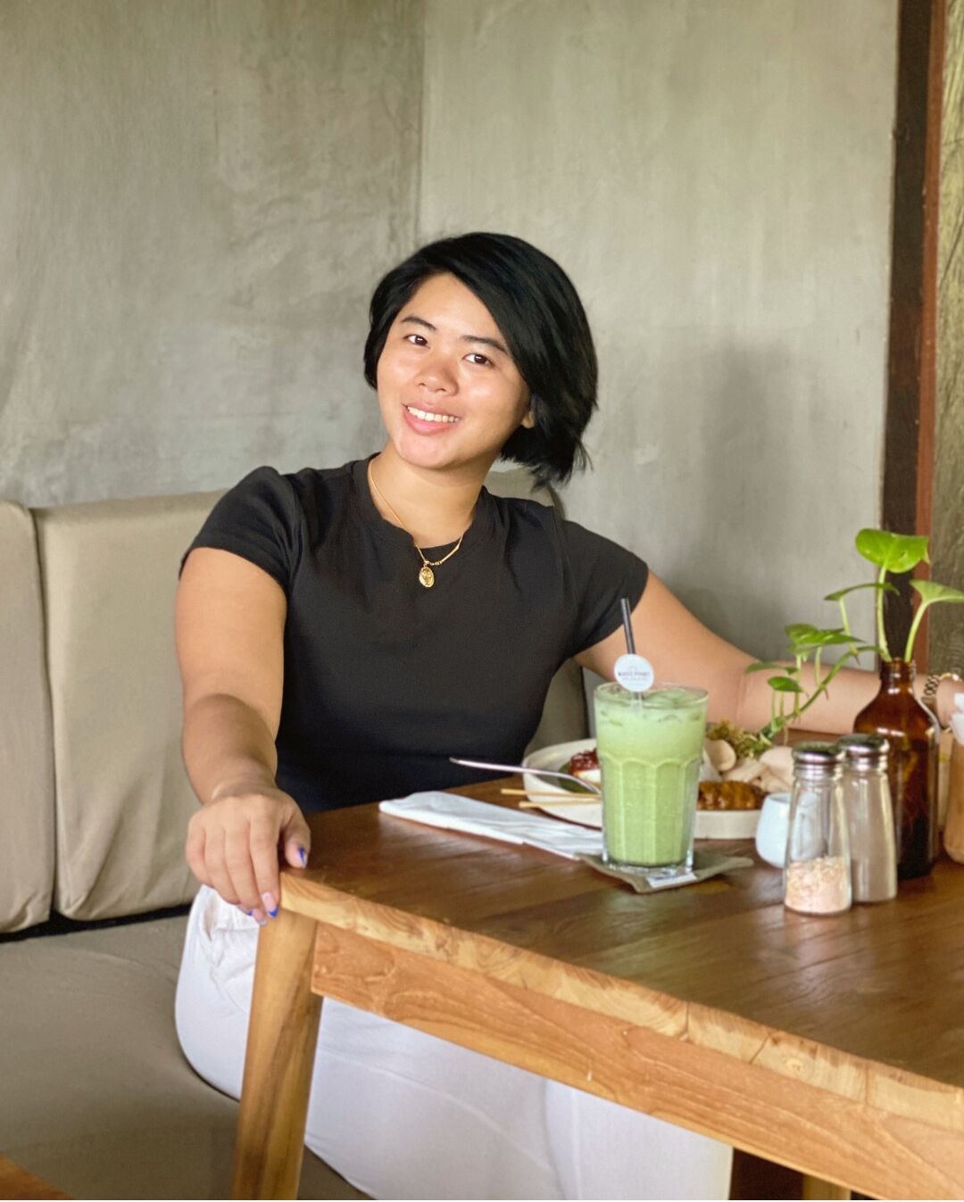 A person seated at a wooden table with a green drink, a plate with food, and salt and pepper shakers.