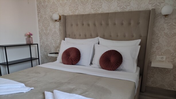 A neatly made bed with white linens and two decorative pillows, a bedside table with a vase, and a wall with a light switch. - Bloom Apartments & Lounge Bar (Vir)