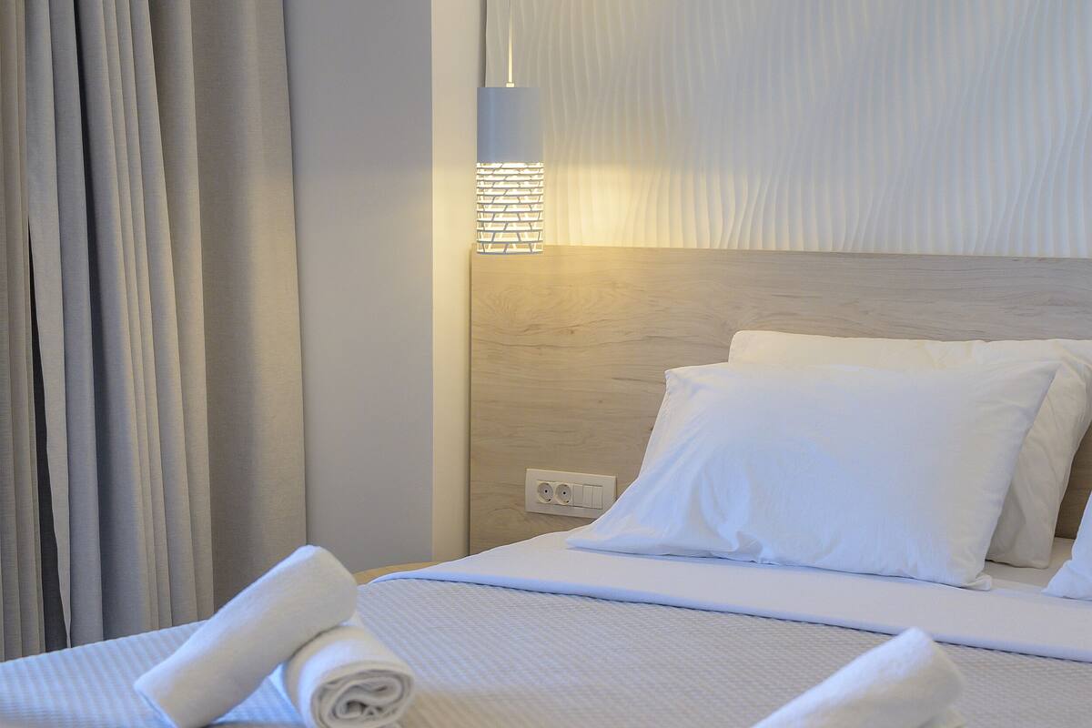 A neatly made bed with white linens and rolled towels, a wooden headboard, and a wall-mounted light fixture.