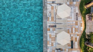 Vista aérea de un área de piscina con piso de baldosas, dos sombrillas y un jardín ajardinado.