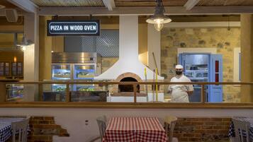 An indoor pizza oven restaurant with a chef, tables with checkered tablecloths, and a sign reading "PIZZA IN WOOD OVEN".