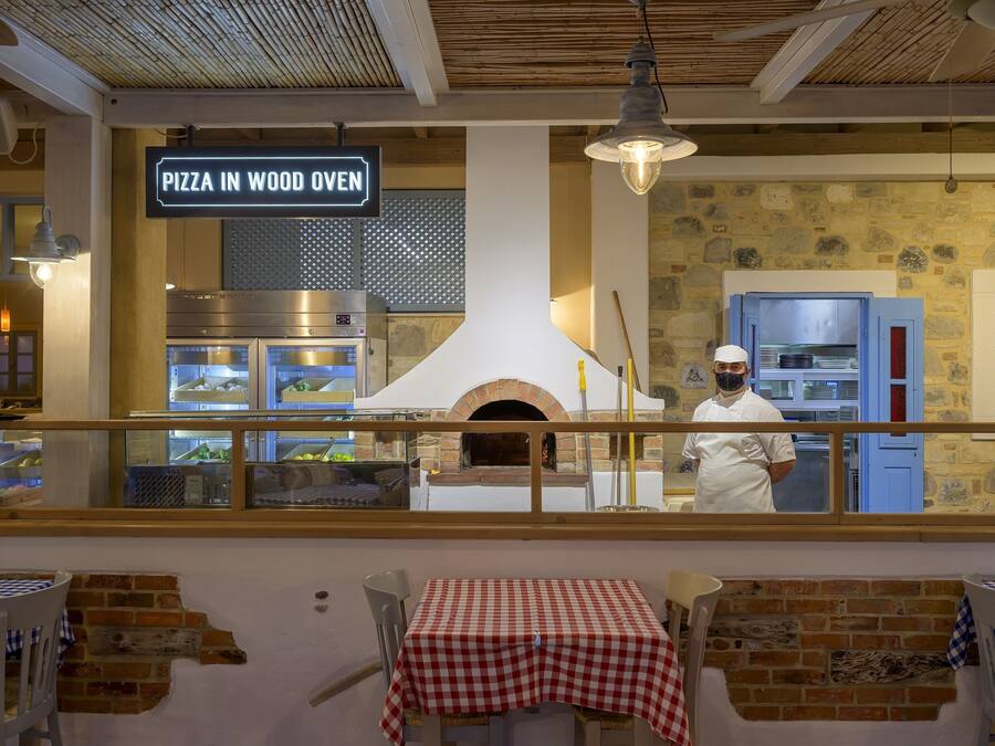 An indoor pizza oven restaurant with a chef, tables with checkered tablecloths, and a sign reading "PIZZA IN WOOD OVEN".