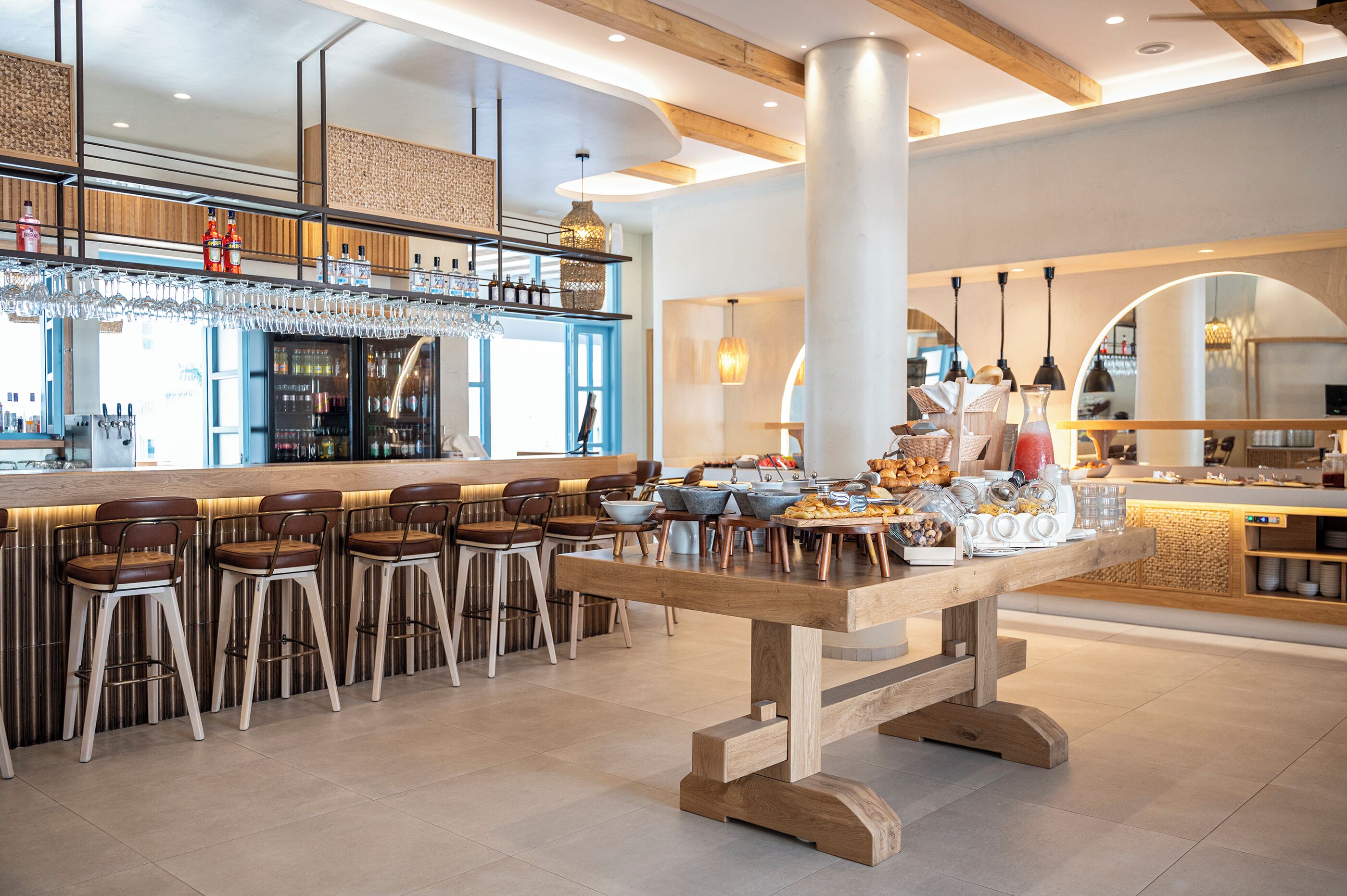 A modern bar area with a long wooden table, bar stools, and a variety of beverages and snacks.
