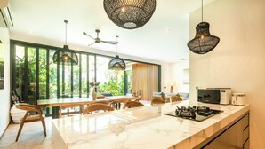 Une cuisine moderne dotée d’un plan de travail en marbre, de luminaires suspendus noirs et offrant une vue sur l’extérieur grâce à de grandes fenêtres.