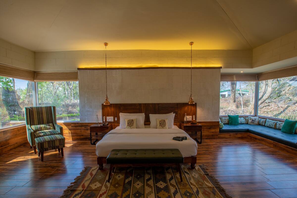 A spacious bedroom with a large bed, a green armchair, a bench, and a view of trees through large windows.