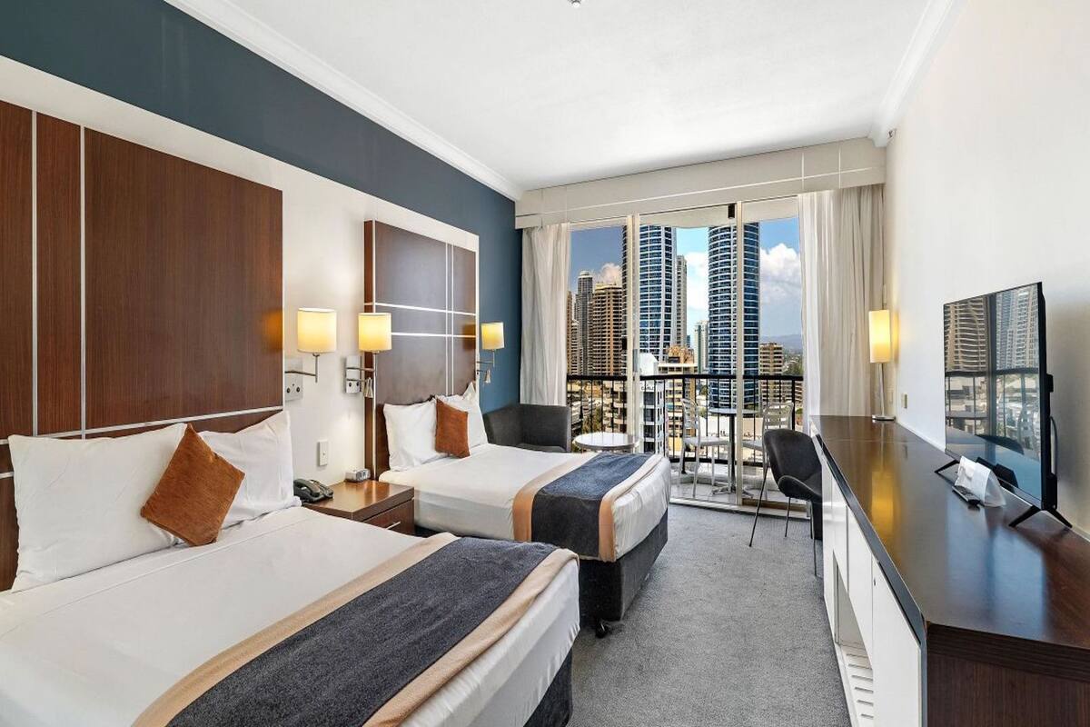 A hotel room with two beds, a desk, and a TV. The room has a city view with tall buildings.