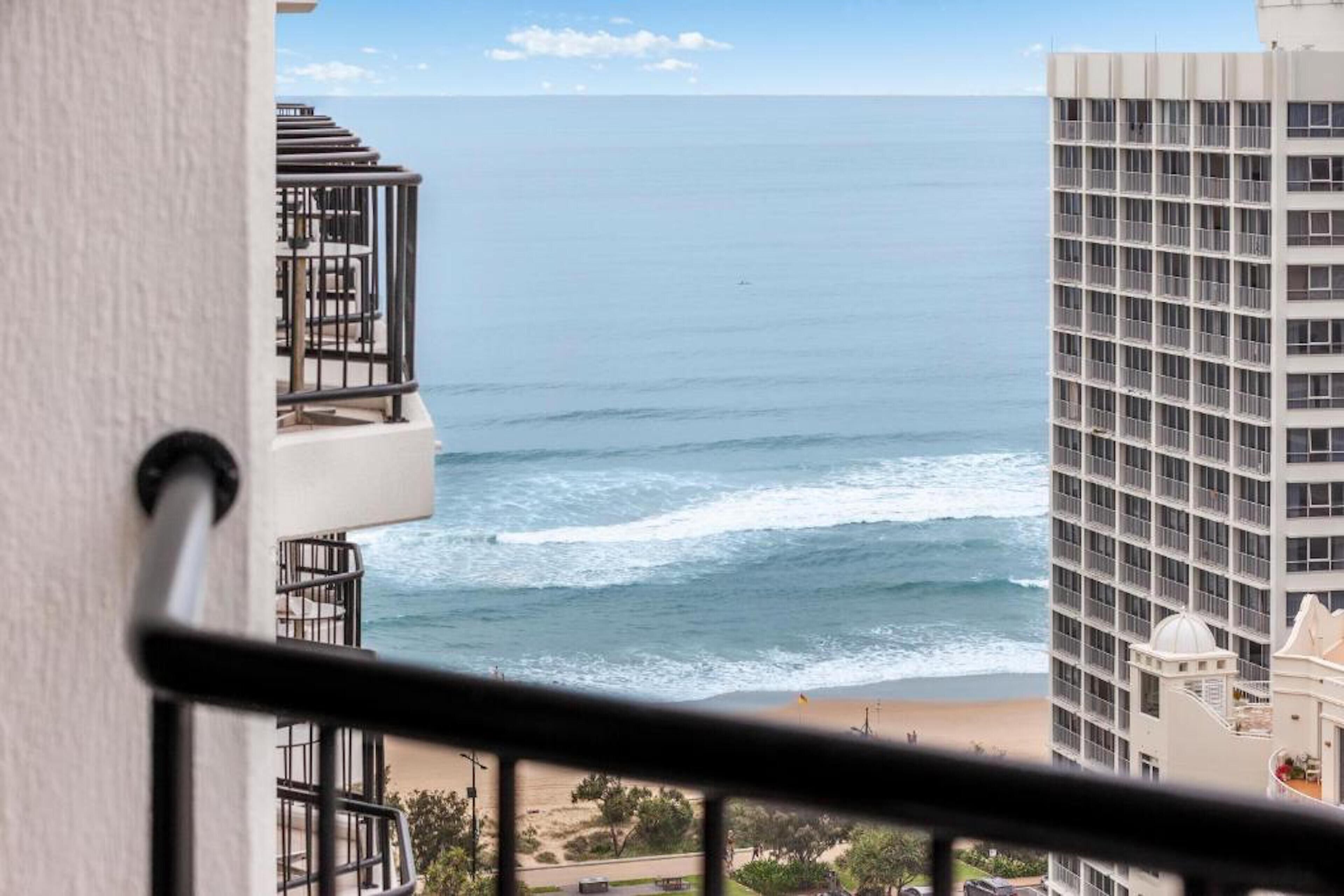 A view of the ocean from a balcony with a railing, a beach, and a tall building in the background.