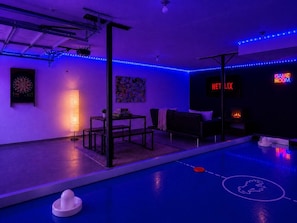 Une pièce comprenant un jeu de fléchettes, une table avec une lampe, un canapé, une cheminée et un panneau néon indiquant qu’il s’agit d’une salle de jeux.