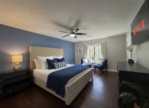 A hotel room with a bed, a desk, a chair, a ceiling fan, and a TV mounted on the wall.