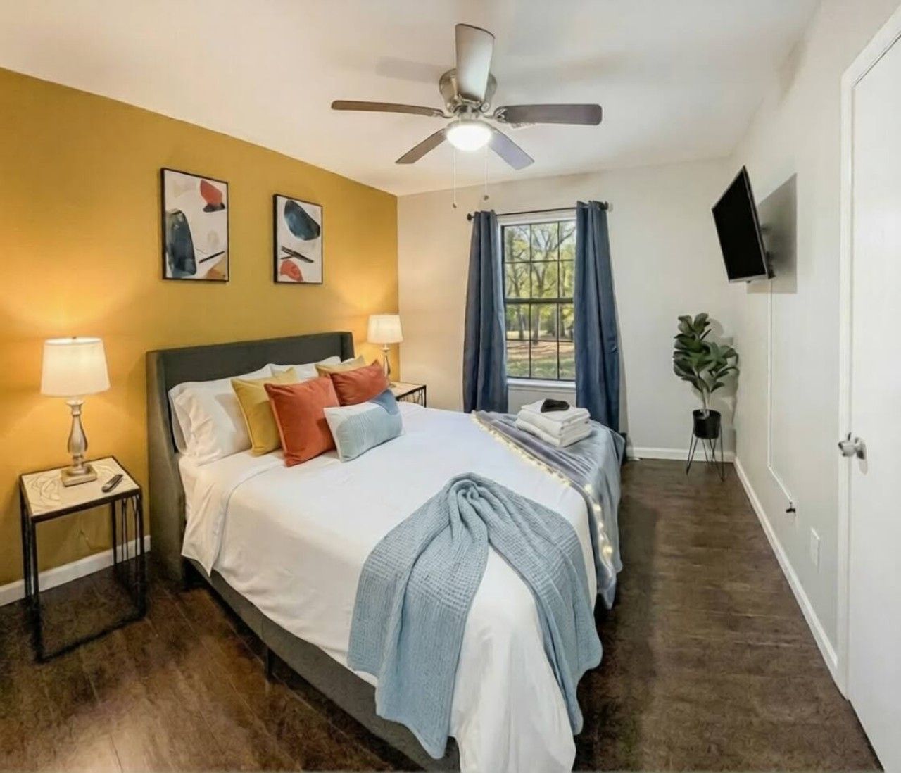 A hotel room with a bed, bedside lamps, a ceiling fan, a window with curtains, a flat-screen TV, and a potted plant.