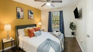 Une chambre d’hôtel comprenant un lit, des lampes de chevet, un ventilateur de plafond, une fenêtre avec des rideaux, un téléviseur à écran plat et une plante en pot.