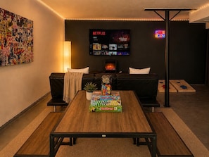 Un salon avec une table basse en bois, deux canapés noirs, un téléviseur diffusant Netflix et un panneau néon indiquant une salle de jeux.