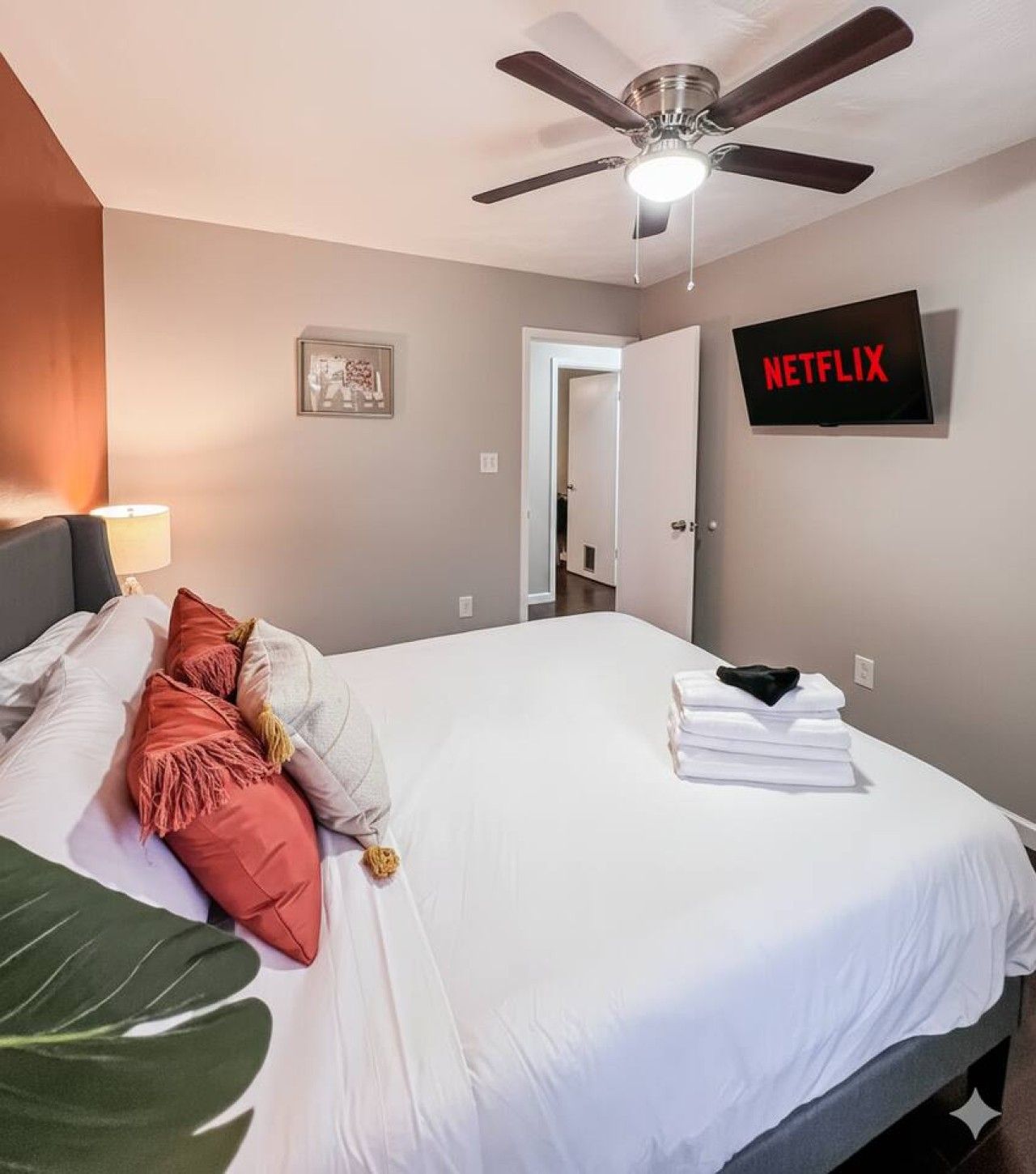 Ein Hotelzimmer mit einem Bett, einem Deckenventilator, einem an der Wand montierten Fernseher mit der Aufschrift 'NETFLIX' und einer Tür zu einem weiteren Raum.