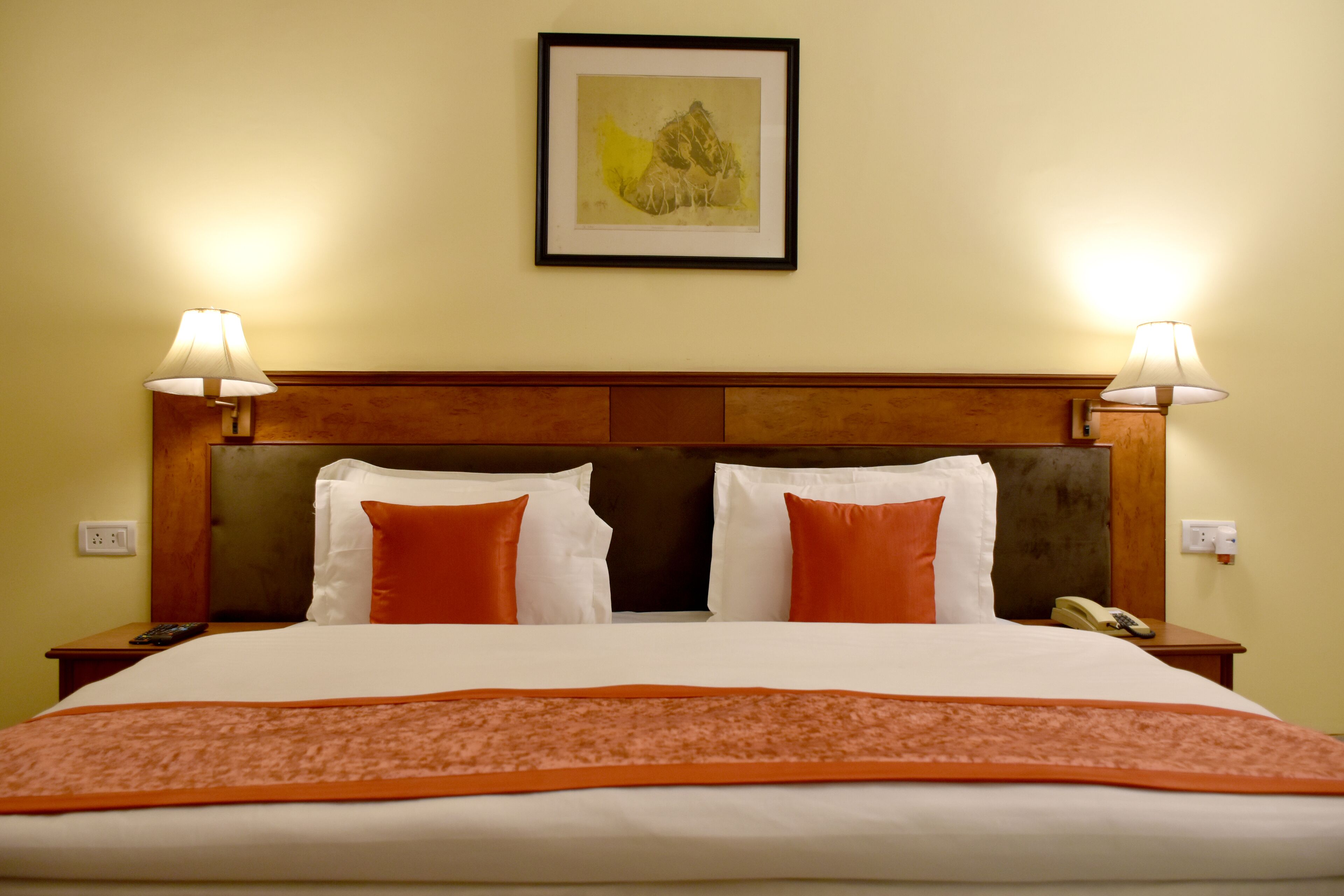 A neatly made bed with white and orange bedding, two bedside lamps, a framed artwork above the bed, and a wooden headboard.