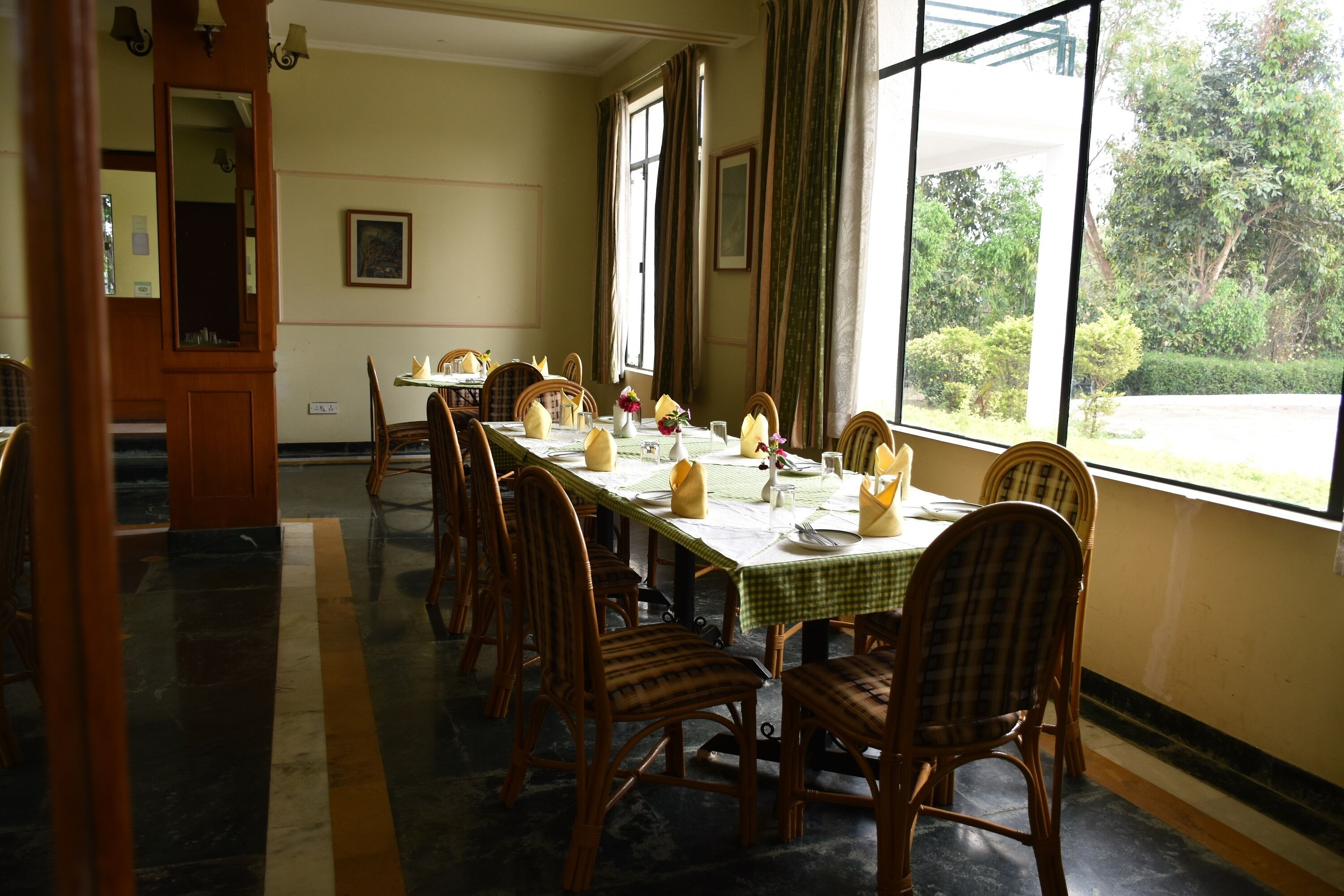 A dining room with a long table set for a meal, surrounded by wicker chairs, with a view of greenery outside.