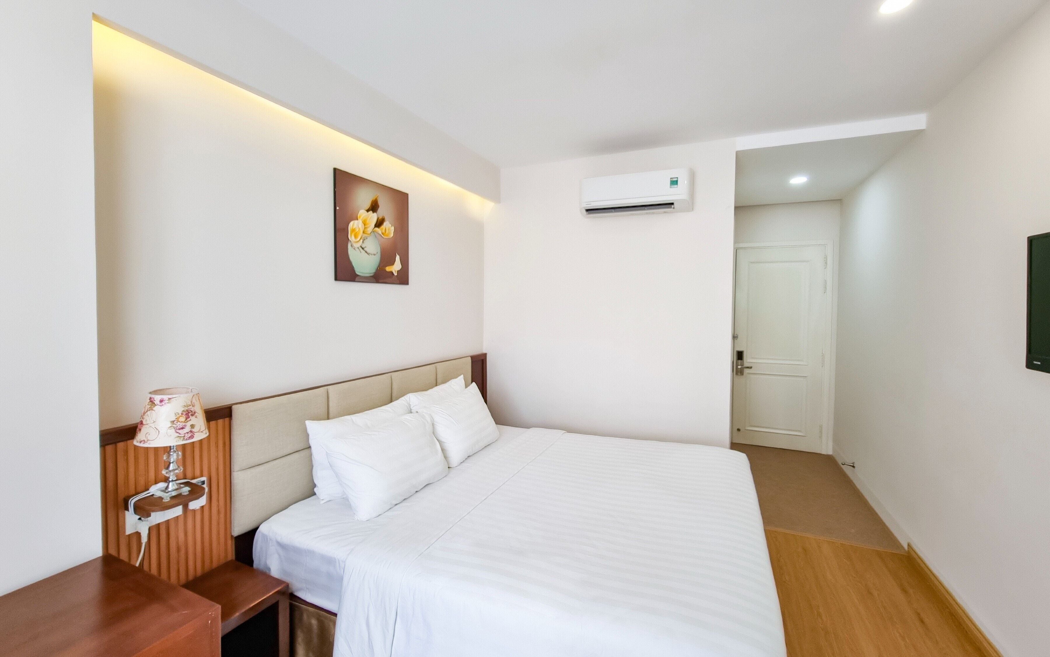 A hotel room with a bed, bedside table, lamp, air conditioning unit, and a painting on the wall.