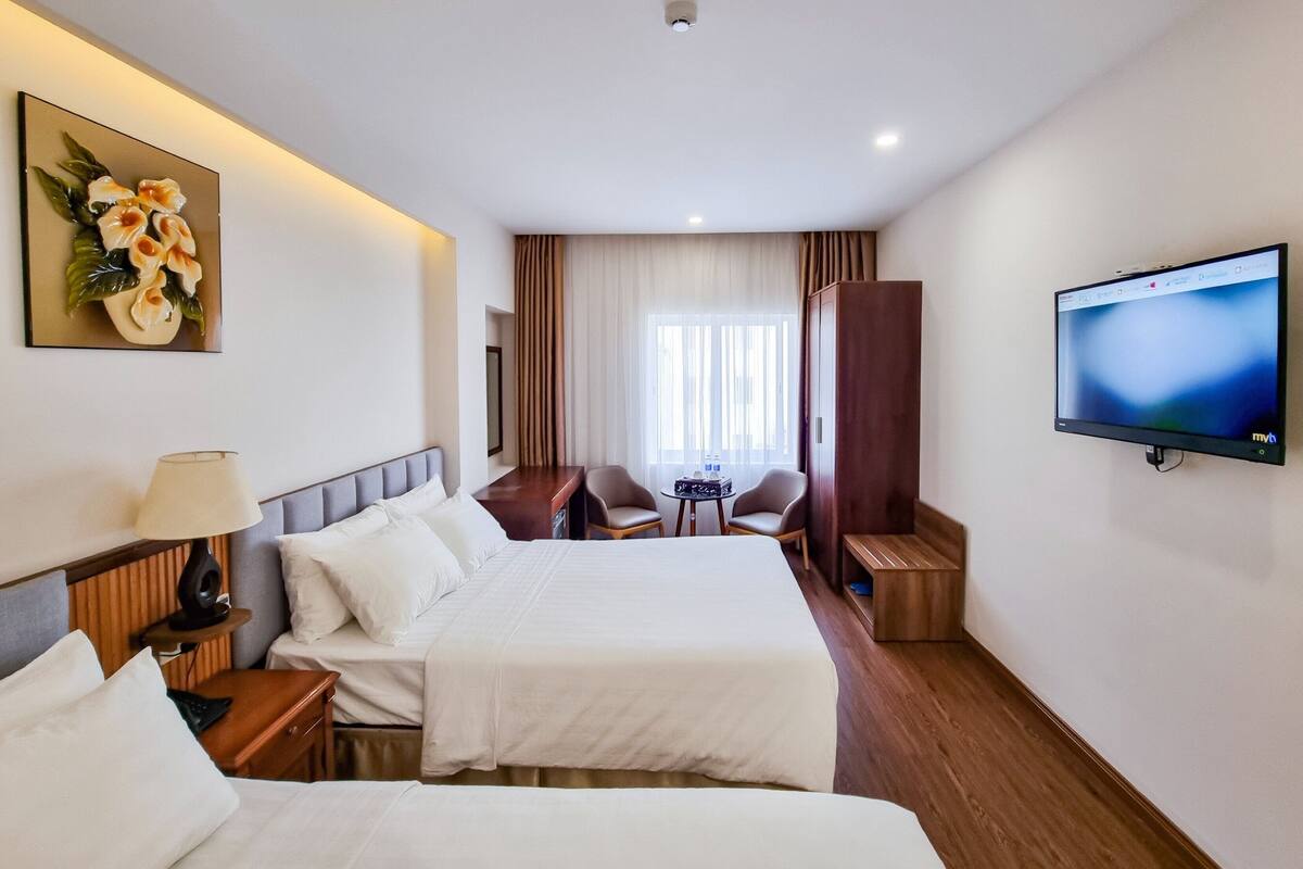 Ein Hotelzimmer mit zwei Betten, einem Schreibtisch, einem Sessel, einem Fernseher und einem Gemälde.