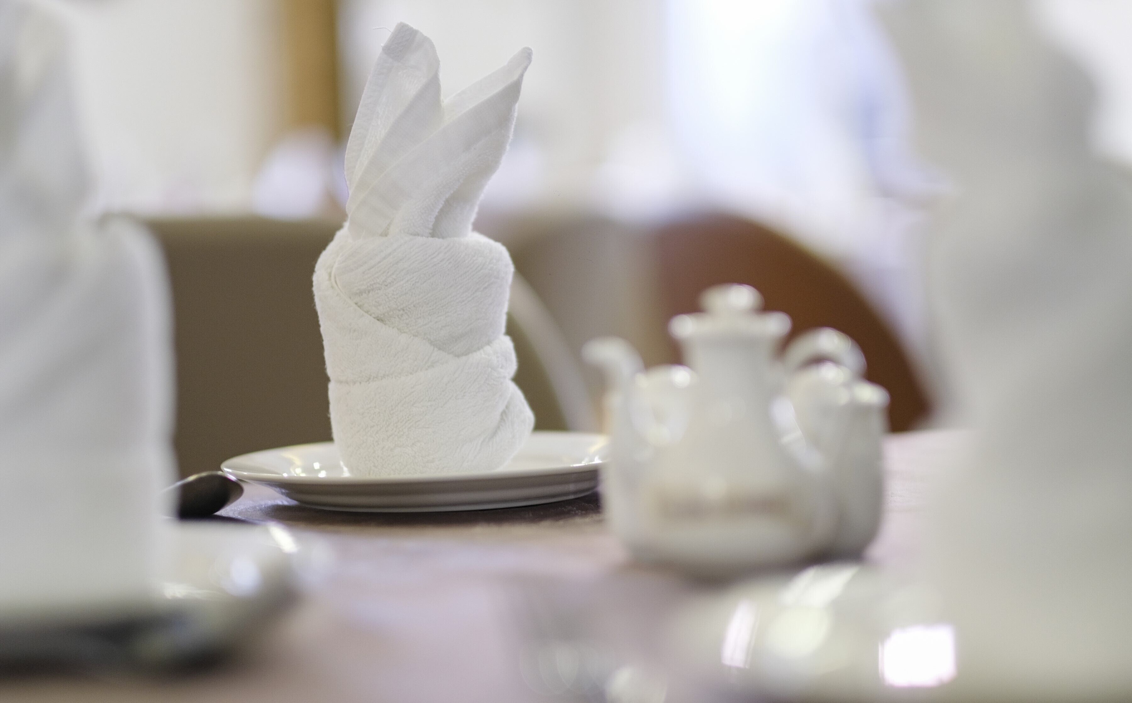 A neatly folded napkin on a plate, a teapot, and a cup on a table.