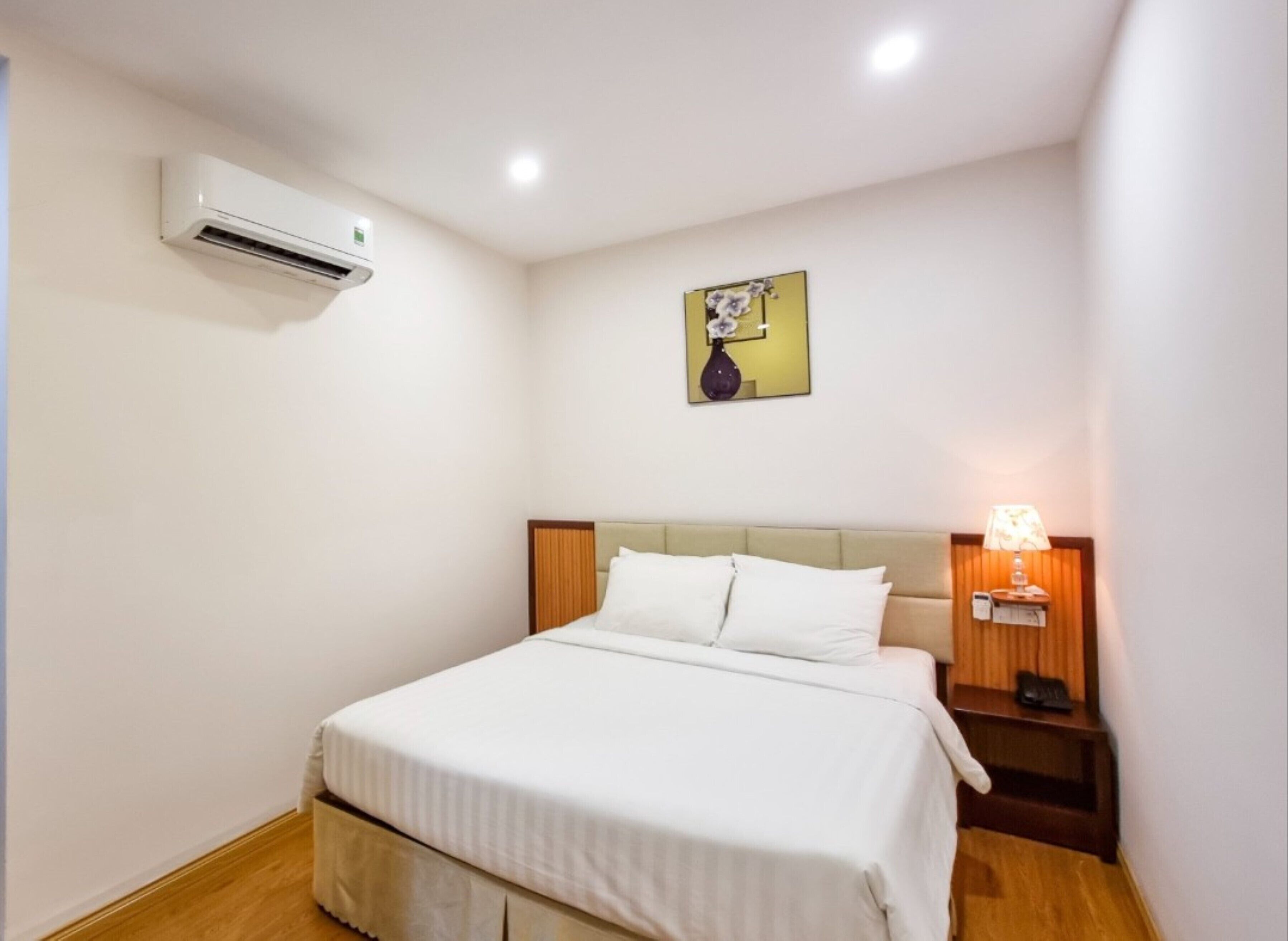 A hotel room with a bed, an air conditioning unit, a bedside lamp, and a painting on the wall.
