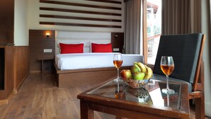 Habitación de hotel con techo de madera, cama con ropa de cama blanca y roja, una mesita de vidrio con fruta y copas de vino, y vista al exterior a través de una ventana con cortinas.