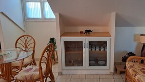Un comedor con mesa de vidrio, sillas de mimbre, un aparador con puertas de cristal y un sofá estampado.