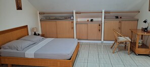Un dormitorio con cama, armario de madera, escritorio, silla y una mesita de noche con lámpara.